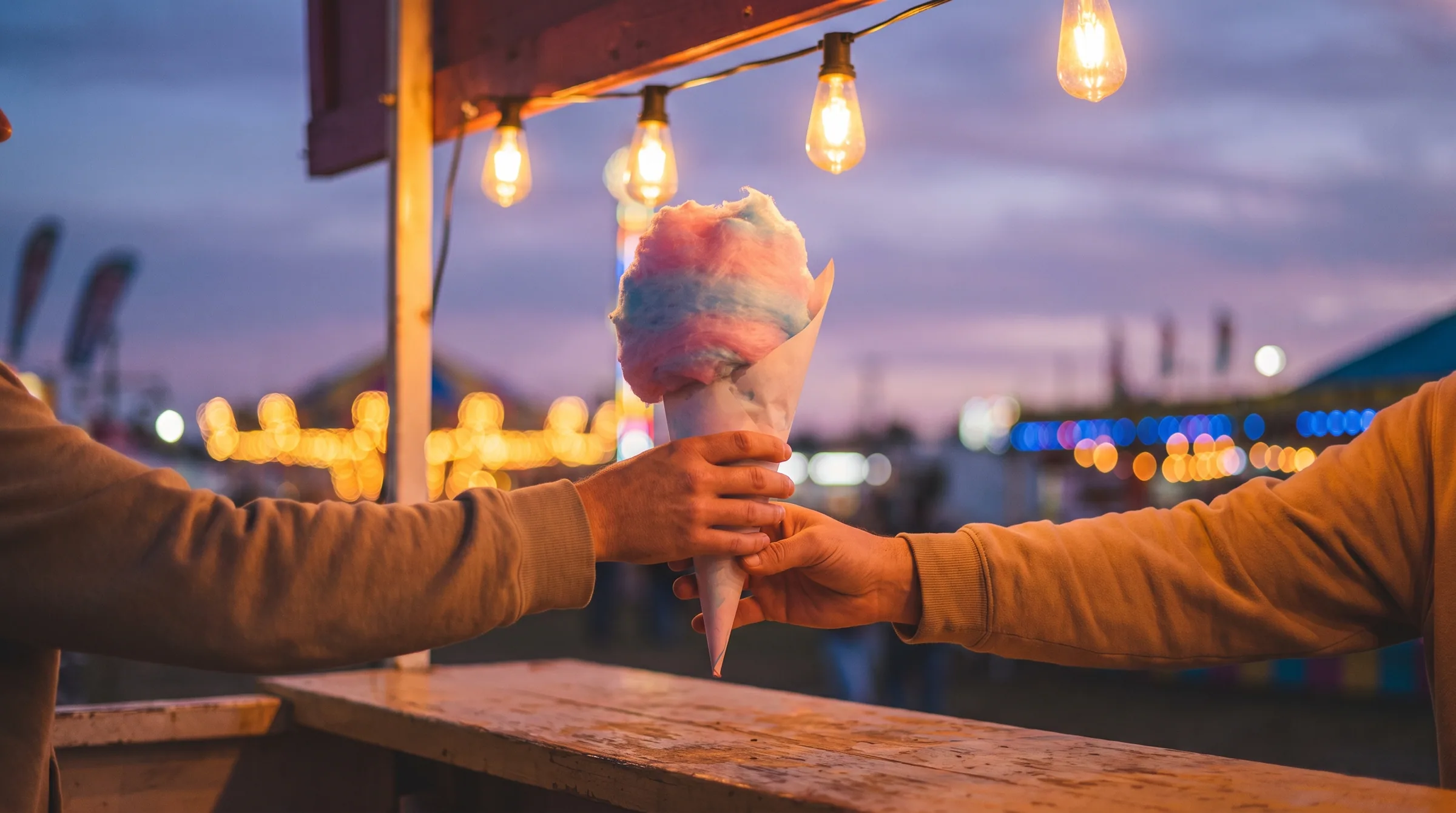 A cone of cotton candy passed between two hands at a lit stand